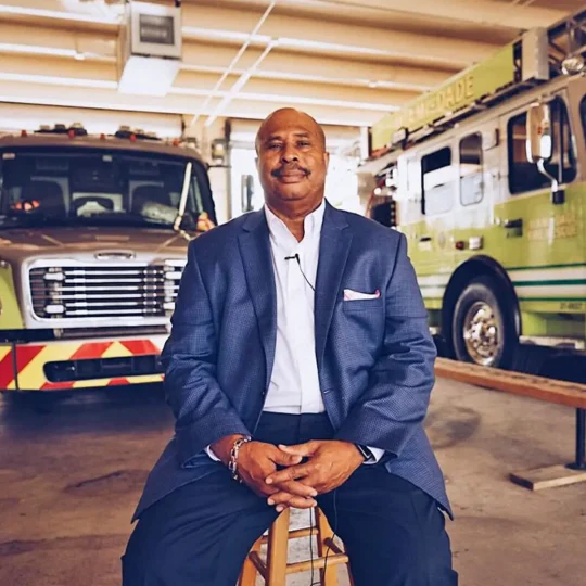 Man with a suit sitting on stool and 2 fire trucks behind the man.